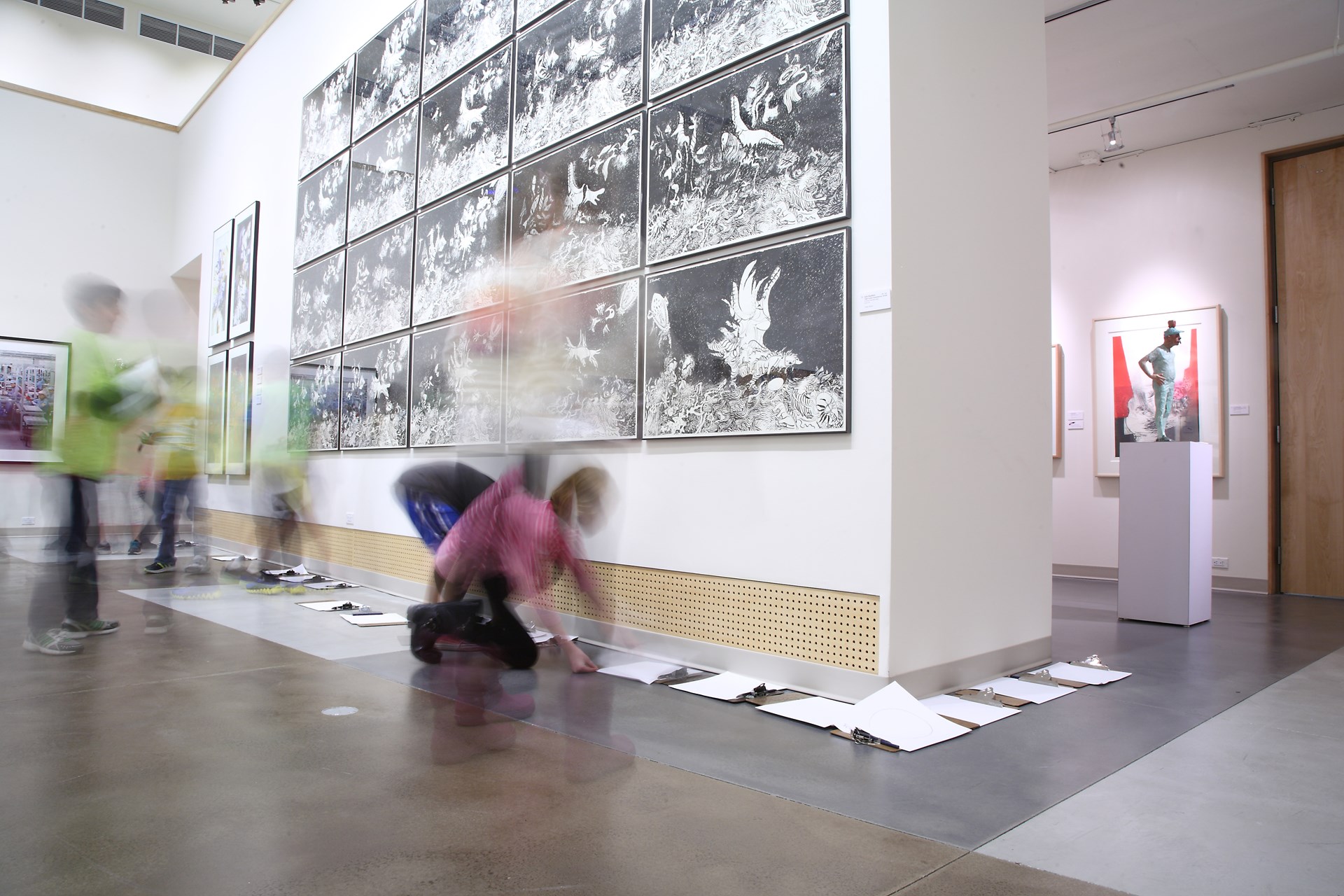 a time-lapse image of six students organizing work on the floor under an exhibit display inside the GS Gallery