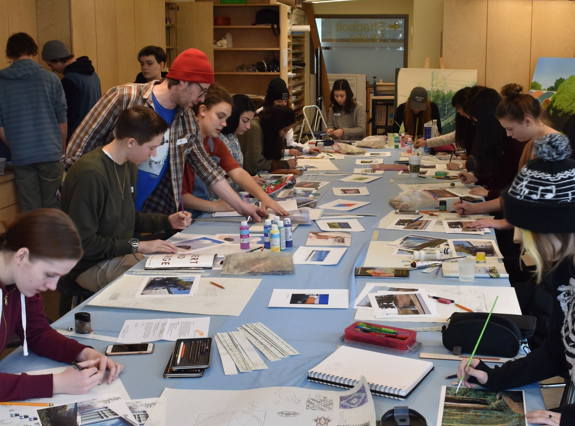 a long table with students working on art projects, with Erin leaning over pointing at something on the table
