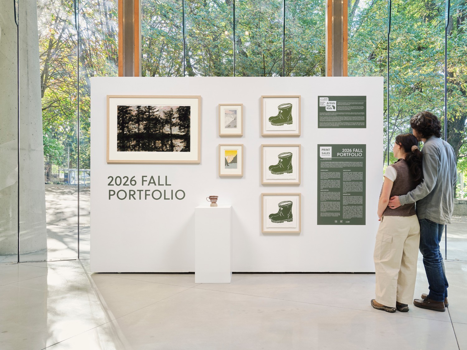 Display of the Artists for Kids 2026 Fall Portfolio in a bright gallery space. Framed artworks and informational panels are mounted on a white exhibition wall, with a ceramic piece on a pedestal in front. Large windows reveal trees outside, and two visitors stand viewing the display.
